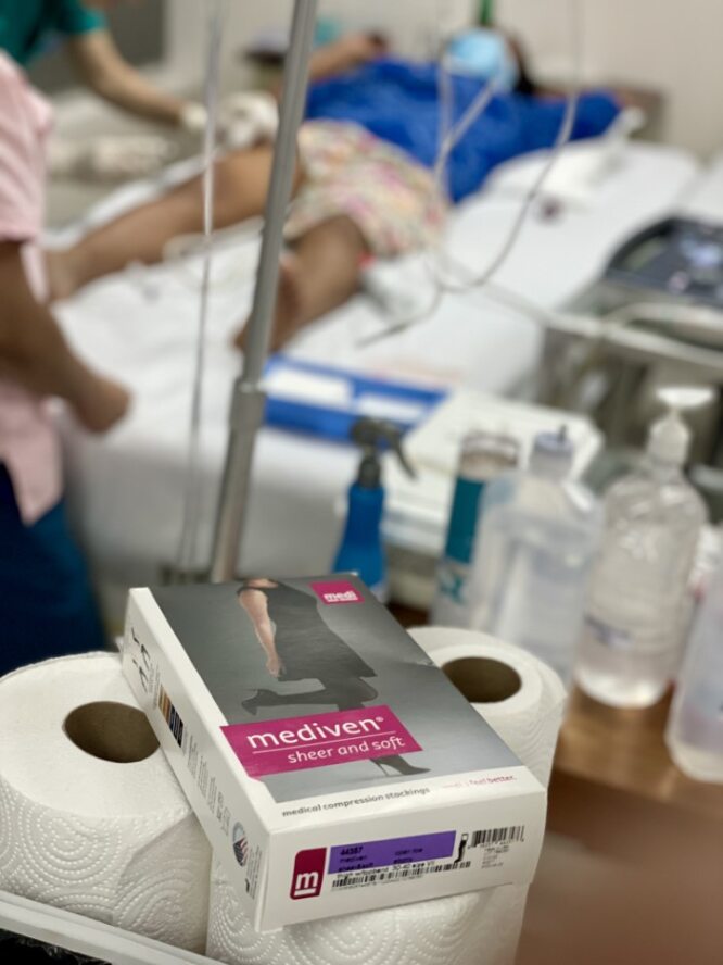 A patient lies on a hospital bed while helpers care for him. In the foreground are medi products for compression stocking.