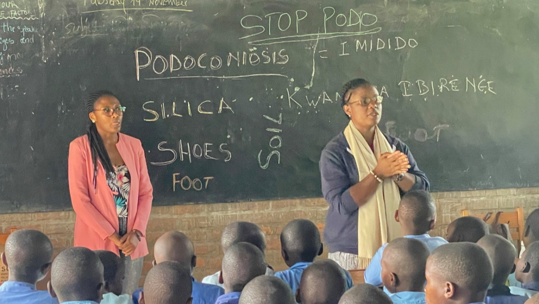 Two teachers stand in front of a blackboard in a school classroom with various terms relating to podoconiosis written on it